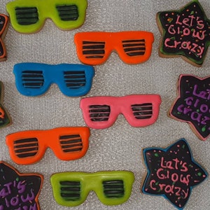 Puede incluir: Nueve galletas con forma de estrella decoradas con glaseado negro y chispas de colores. Las galletas tienen el texto "Let's Glow Crazy" escrito en ellas. Algunas galletas también tienen gafas de sol de colores dibujadas en ellas.