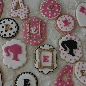 Puede incluir: Una colección de galletas de azúcar decoradas con glaseado rosa, blanco, negro y dorado. Las galletas tienen forma de corazones, números y marcos. Algunas galletas tienen el nombre "Evie" escrito en ellas, mientras que otras tienen el número "4" en ellas.