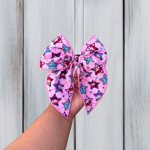 May include: A large hair bow featuring a light pink fabric base with a repeating pattern of blue and purple stars. Each star contains a small illustrated character face and the text "HUNTRIX". The bow edges are finished with bright pink piping.