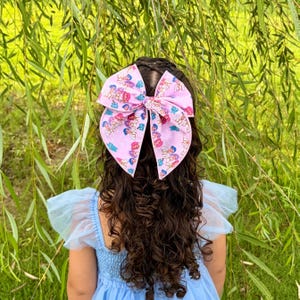 May include: A large hair bow in light pink fabric featuring a repeating pattern of cartoon characters with blue, purple, and orange hair. The bow is tied in a classic style with two long, wide loops and two trailing ends, secured to dark curly hair.