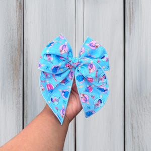 May include: A light blue hair bow featuring a repeating pattern of various cartoon characters enclosed within colorful Christmas ornament shapes, interspersed with small red and white candy canes and tiny white star-like accents.
