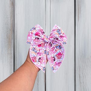 May include: A hair bow featuring a repeating pattern of cartoon characters and stars in shades of pink, blue, and white. The bow has a structured shape with two large loops and two pointed tails, finished with a light pink stitched edge trim.