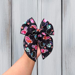 May include: A black hair bow featuring a repeating pattern of cartoon characters, cauldrons, and sparkling disco balls. The fabric has a dark background with small white star-like dots scattered throughout, and the bow is finished with black edges.