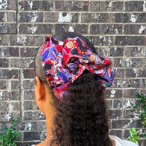 May include: A large fabric hair scrunchie featuring a vibrant, multi-colored graphic print with cartoon-style characters, abstract shapes, and floral motifs in shades of purple, red, pink, and yellow, worn around a ponytail.
