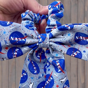 May include: A gray scrunchie with a bow featuring a NASA logo pattern. The design includes blue NASA logos, white stars, and red accents on a gray background. The scrunchie is held by a hand.