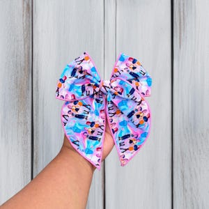 May include: A hair bow featuring a repeating pattern of cartoon characters on a light blue and pink tie-dye background. The bow is constructed from fabric with a pink stitched edge, showcasing multiple figures in various poses across the surface.
