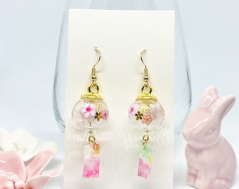 May include: A pair of gold earrings with clear glass charms featuring pink and white flowers. The charms are attached to a small pink and white patterned ribbon.