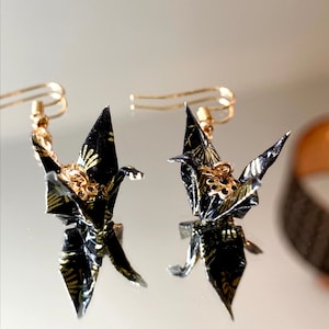 以下が含まれることがあります： 黒と金の模様の紙でできた、金の折り鶴のイヤリングのペア。鶴は、小さな金のビーズが付いた金のチェーンからぶら下がっています。