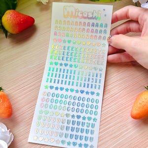 May include: A sheet of holographic stickers with the word "Micashi" in rainbow lettering. The stickers include the alphabet, numbers, hearts, stars, and other shapes in various colors.