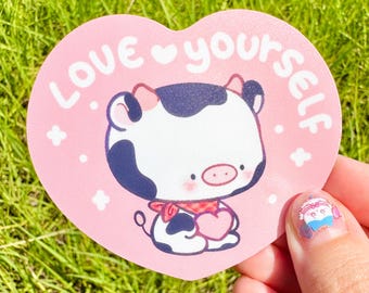 May include: A pink heart-shaped sticker with the words "Love yourself" in white. The sticker features a cartoon cow holding a heart. The cow is black and white with a red scarf. The background is green grass.