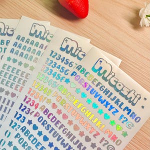 May include: Sheets of holographic stickers with letters, numbers, and symbols in various colors. The stickers include the word "Micashi" in a bubble font. A red and yellow strawberry sits above the stickers.
