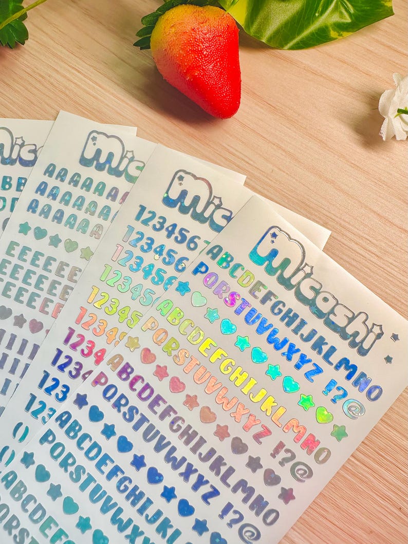 May include: Sheets of holographic stickers with letters, numbers, and symbols in various colors. The stickers include the word "Micashi" in a bubble font. A red and yellow strawberry sits above the stickers.
