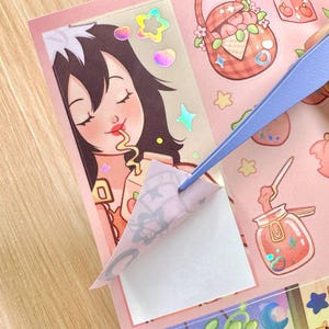 May include: A sheet of pink stickers with various illustrations, including a girl eating, a picnic basket, and a jar of jam. A pair of blue tweezers is peeling off a sticker. The stickers have a holographic effect.