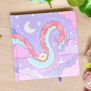 May include: A square notebook with a light purple cover featuring a long, winding light blue dragon with a pink mane and a small yellow chick resting on its body. The background includes pink clouds, a white crescent moon, and white stars.