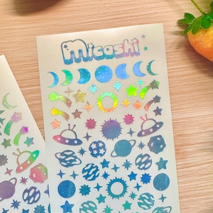 May include: Holographic stickers featuring celestial and space-themed designs. The stickers include moons, stars, planets, shooting stars, suns, and UFOs. The word "Micoshi" is printed at the top of the sticker sheet in a bubble font.