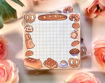May include: A set of 4x6 inch gridded memo pads with a bakery theme. The memo pads have a white background with a grid pattern and are surrounded by a border of various bakery items, including bread, pastries, and coffee.