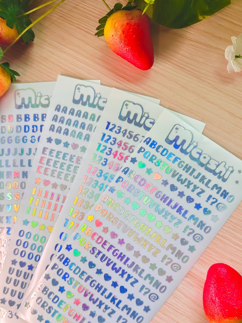 May include: Sheets of holographic alphabet and number stickers with hearts, stars, and other symbols. The stickers have the brand name "Micashi" printed on them. Strawberries are in the background.