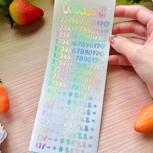 May include: A sheet of iridescent stickers with the name "Micashi" at the top. The stickers feature numbers, symbols, and punctuation marks in various colors. The sheet is held by a hand, with a blurred background.