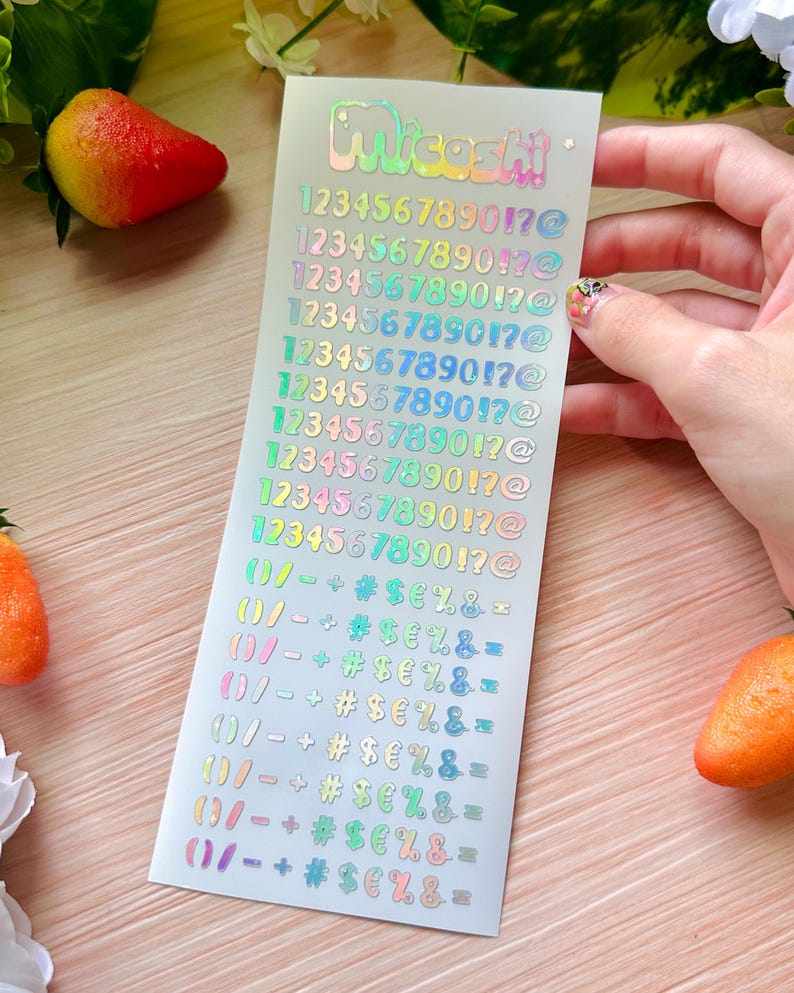 May include: A sheet of iridescent stickers with the name "Micashi" at the top. The stickers feature numbers, symbols, and punctuation marks in various colors. The sheet is held by a hand, with a blurred background.