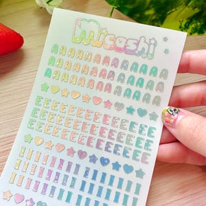 May include: A sheet of iridescent stickers with the word "Micoshi" at the top. The sheet contains multiple rows of the letter "A", "E", "I", and the numbers "0", along with star and heart shapes, all in a rainbow color scheme.
