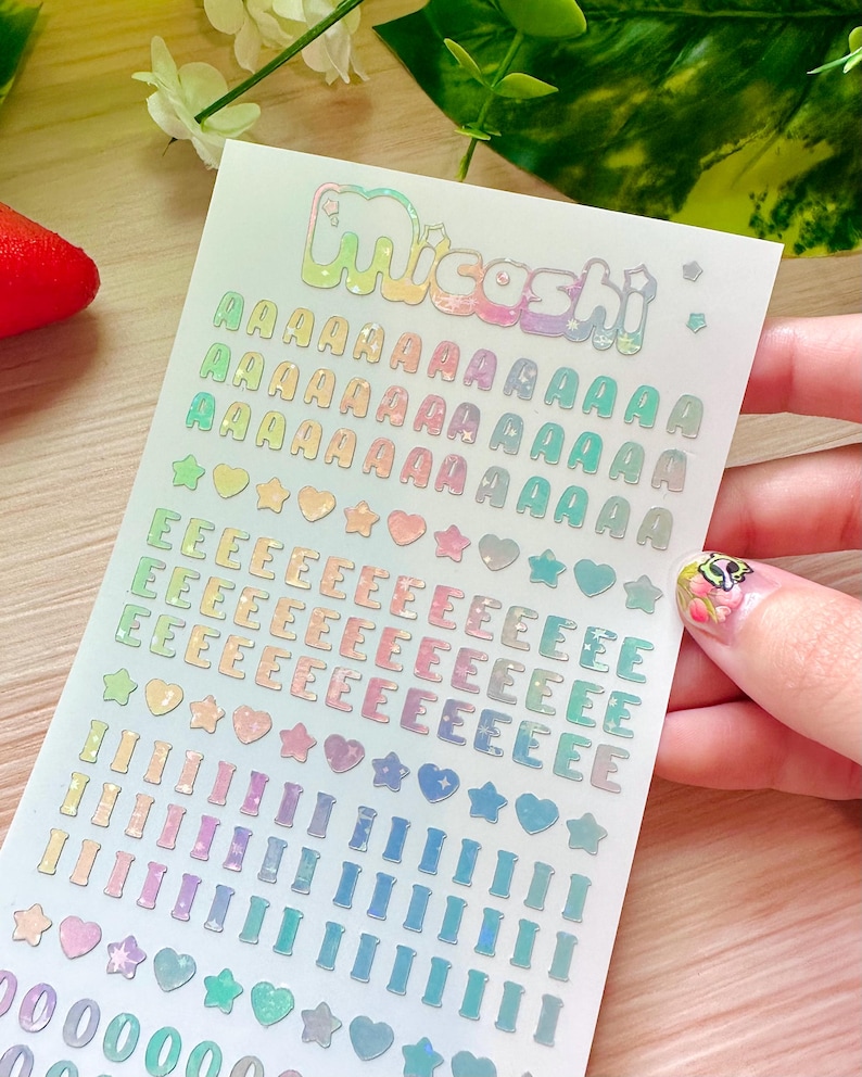 May include: A sheet of iridescent stickers with the word "Micoshi" at the top. The sheet contains multiple rows of the letter "A", "E", "I", and the numbers "0", along with star and heart shapes, all in a rainbow color scheme.