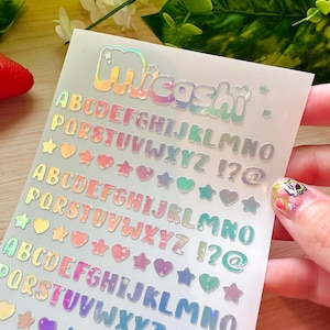 May include: Holographic alphabet stickers with the word "Micashi" at the top. The stickers include the alphabet, punctuation marks, and heart and star shapes in various colors. The stickers are held by a person.
