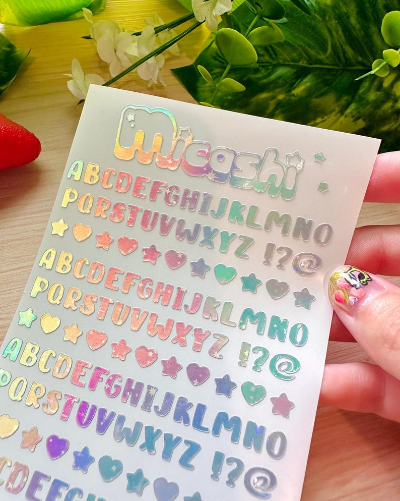 May include: Holographic alphabet stickers with the word "Micashi" at the top. The stickers include the alphabet, punctuation marks, and heart and star shapes in various colors. The stickers are held by a person.