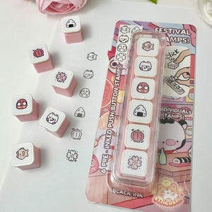 May include: A set of six square pre-inked push button stamps with pink bases and white tops featuring black line art icons of a fish, cat, daruma, onigiri, lantern, and cherry blossom. The packaging reads FESTIVAL STAMPS and BLACK INK.