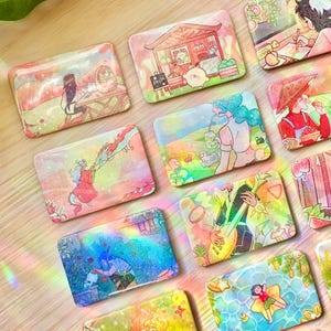 Puede incluir: Una colección de piezas de arte de resina rectangulares y coloridas con varias ilustraciones. La obra de arte presenta escenas con personas, animales y paisajes en un estilo de dibujos animados vibrante. Las piezas están sobre una superficie de madera clara.