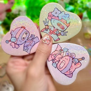 May include: Three heart-shaped compact mirrors with cartoon character designs. The mirrors have a holographic, glittery finish and feature characters in shades of pink, purple, and yellow. Each character has a unique design, with stars and other embellishments.