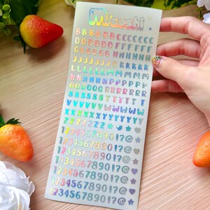 May include: A sheet of holographic stickers with the word "Miceshi" at the top. The stickers include the alphabet, numbers, and symbols in various colors. The stickers are on a white background.