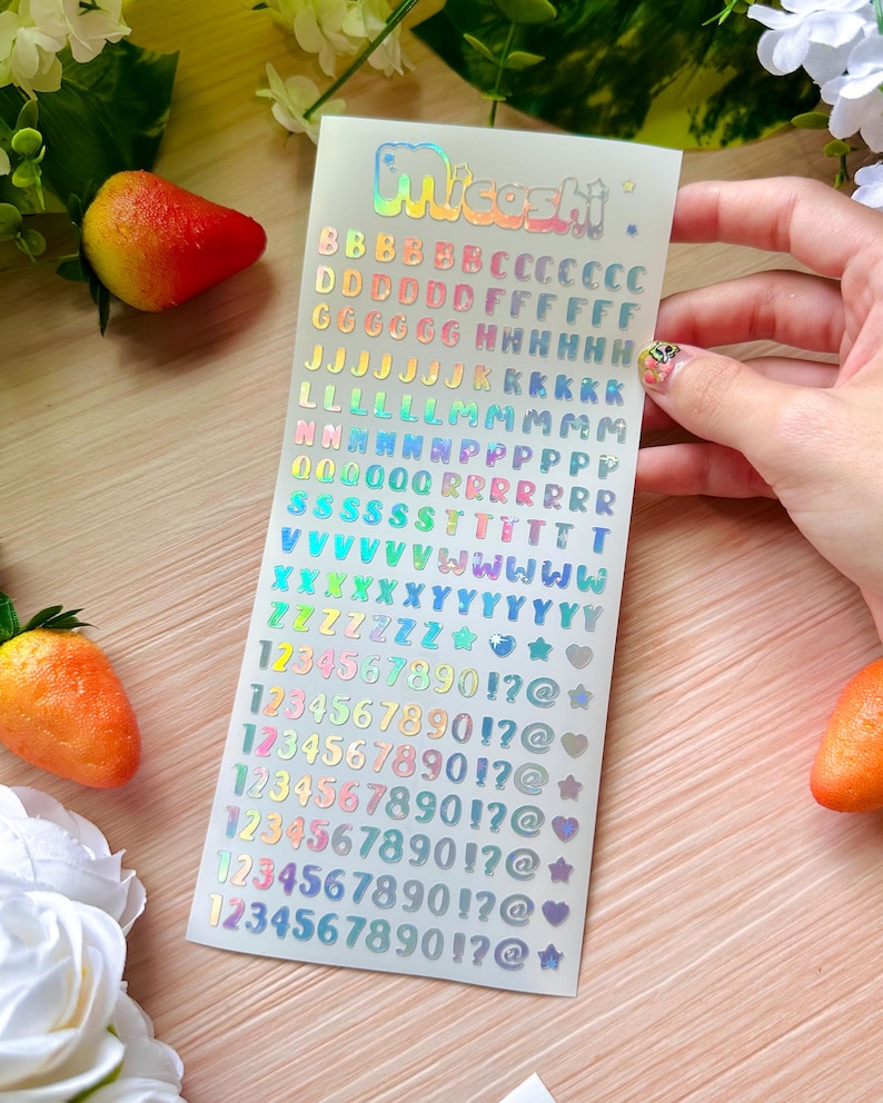 May include: A sheet of holographic stickers with the word "Miceshi" at the top. The stickers include the alphabet, numbers, and symbols in various colors. The stickers are on a white background.