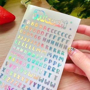 May include: A sheet of iridescent stickers with the word "Miceshi" at the top. The sheet contains colorful alphabet letters, numbers, stars, and hearts. The stickers are in various colors, including pink, blue, green, and yellow.