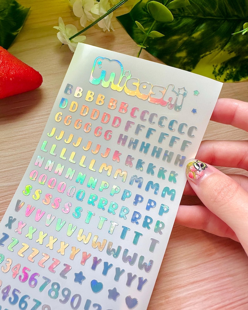 May include: A sheet of iridescent stickers with the word "Miceshi" at the top. The sheet contains colorful alphabet letters, numbers, stars, and hearts. The stickers are in various colors, including pink, blue, green, and yellow.