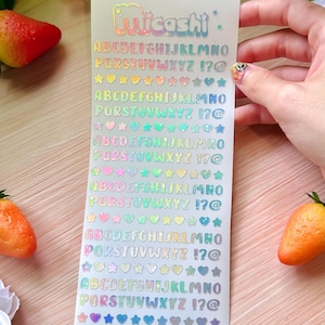 May include: A sheet of holographic stickers with the brand name "Micoshi" at the top. The stickers feature the alphabet, numbers, and symbols in a rainbow color scheme. Various shapes like hearts and stars are also included.