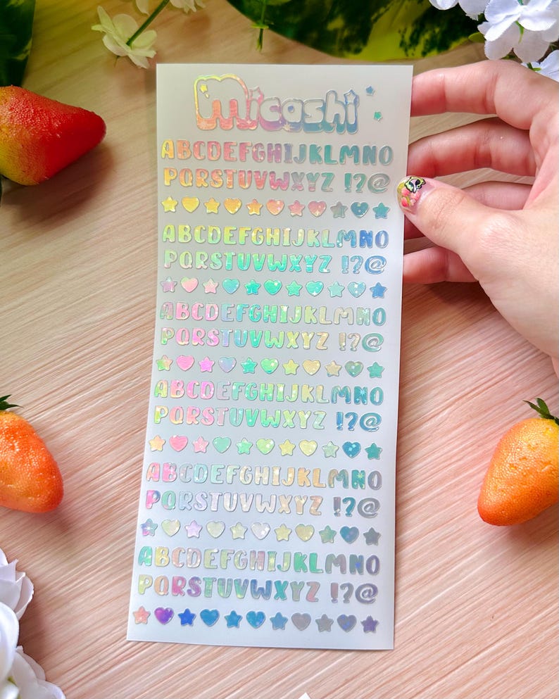 May include: A sheet of holographic stickers with the brand name "Micoshi" at the top. The stickers feature the alphabet, numbers, and symbols in a rainbow color scheme. Various shapes like hearts and stars are also included.