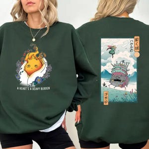 Op de afbeelding: Donkergroene sweatshirt met een witte kraag. De voorkant heeft een afbeelding van een vlamachtig wezen in een hand, met de tekst "A HEART'S A HEAVY BURDEN". De achterkant heeft een Japanse anime-stijl illustratie.