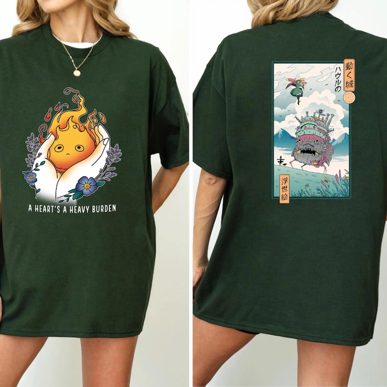 Op de afbeelding: Donkergroen t-shirt met een afbeelding van een cartoon vuurgeest in handen, met de tekst "A Heart's A Heavy Burden." De achterkant heeft een Japanse illustratie van een kasteel en personages.