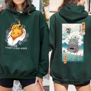 Op de afbeelding: Donkergroene hoodie met een grafische afbeelding aan de voorkant van een cartoon vuurgeest in een hand, met de tekst "A HEART'S A HEAVY BURDEN". De achterkant heeft een illustratie in Japanse stijl met tekst.