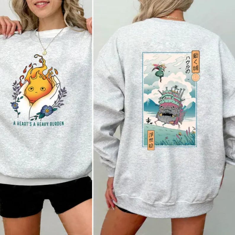 Op de afbeelding: Een lichtgrijs sweatshirt met twee ontwerpen. De voorkant toont een cartoon vuurgeest in handen, met de tekst "A HEART'S A HEAVY BURDEN". De achterkant toont een Japanse illustratie van een bewegend kasteel en personages.
