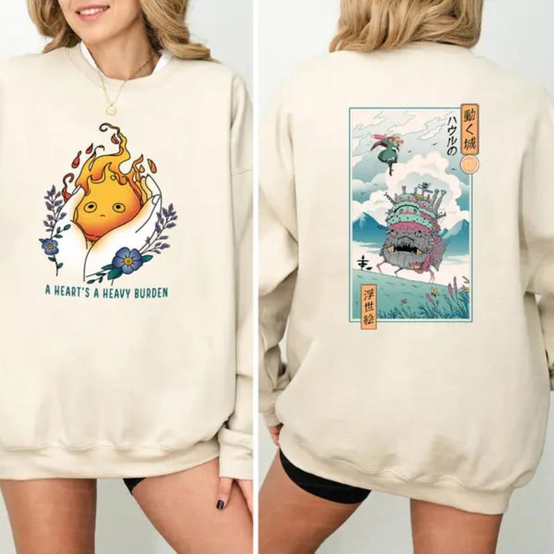 Op de afbeelding: Cr&egrave;mekleurige sweatshirt met een afbeelding aan de voorkant van een vlamachtig wezen dat in een hand wordt gehouden, omringd door bloemen, met de tekst "A HEART'S A HEAVY BURDEN." De achterkant toont een Japanse illustratie van een bewegend kasteel en tekst.