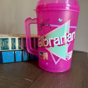 May include: Pink plastic tumbler with a handle and a straw. The tumbler has a design of glasses, stars, and books with the word "Librarian" in a colorful triangle.
