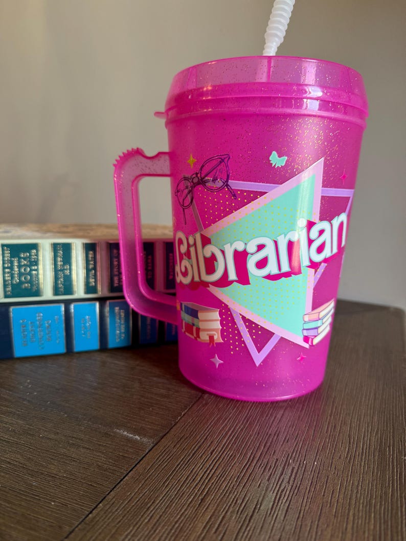 May include: Pink plastic tumbler with a handle and a straw. The tumbler has a design of glasses, stars, and books with the word "Librarian" in a colorful triangle.