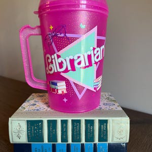 May include: Pink glitter tumbler with a handle and a straw. The tumbler has a turquoise triangle with the word "Librarian" in pink letters. There are images of glasses, books, and butterflies on the tumbler.