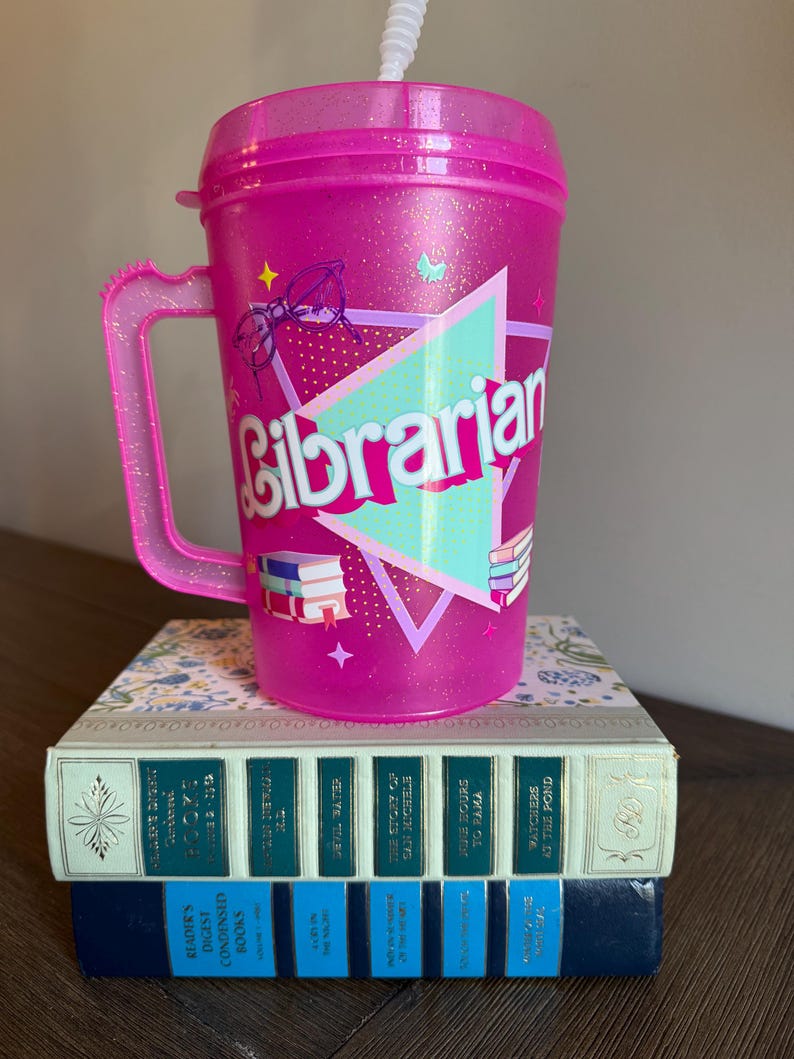 May include: Pink glitter tumbler with a handle and a straw. The tumbler has a turquoise triangle with the word "Librarian" in pink letters. There are images of glasses, books, and butterflies on the tumbler.