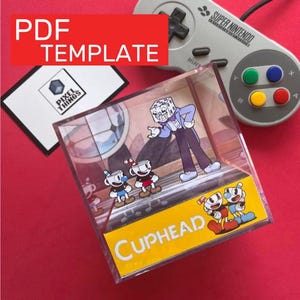 Pode incluir: Uma caixa de exibição de acrílico transparente com personagens de Cuphead e o texto "Cuphead" em amarelo. Um controle Super Nintendo e um modelo PDF também são visíveis.