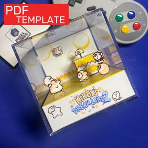 Peut inclure: Un présentoir en acrylique transparent avec un diorama en pixel art de Kirby's Dream Land 2. Le diorama comprend des personnages et des décors pixélisés, avec le texte "Kirby's Dream Land 2" et "PDF TEMPLATE" visible. Une manette de jeu rétro est en arrière-plan.