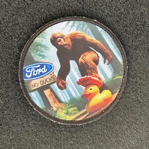 Può includere: Toppa rotonda con un'illustrazione di Bigfoot che calpesta una paperella di gomma. La toppa include il logo Ford e il testo "NO DUCKS" su un cartello. Lo sfondo mostra una scena di foresta con alberi e cielo blu.