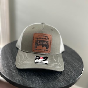 May include: A gray and white trucker hat with a brown leather patch featuring a black and white illustration of a Ford Bronco. The patch has the text "Bronco" in white lettering. The hat has a black and red Richardson label with the text "Richardson" and "112" in white lettering.