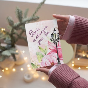 Pu&ograve; includere: Un biglietto di auguri con la scritta "Paris is always a good idea" in corsivo viola. Il biglietto presenta un'illustrazione ad acquerello di un lampione e fiori rosa. Il biglietto &egrave; tenuto da una persona che indossa un maglione rosa.